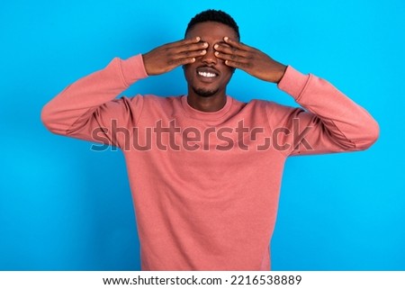 young handsome man wearing pink sweater over blue background covering eyes with hands smiling cheerful and funny. Blind concept. Royalty-Free Stock Photo #2216538889