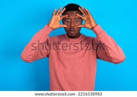young handsome man wearing pink sweater over blue background keeping eyes opened to find a success opportunity.