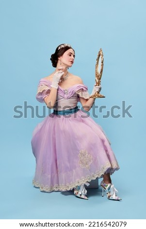 Young and pretty. Beautiful charming woman in lilac color vintage dress as a royal person, princess looking in mirror at herself on blue background. Concept of comparison of eras, renaissance style Royalty-Free Stock Photo #2216542079