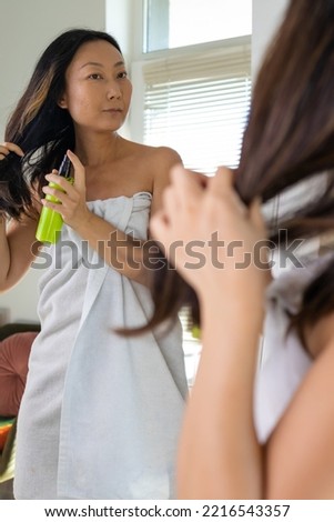 Haircare. Smiling Asian Woman Applying Hair Spray To Split Ends, Young Happy Female Standing Wrapped In Towel After Shower Using avocado conditioner At Home. Royalty-Free Stock Photo #2216543357