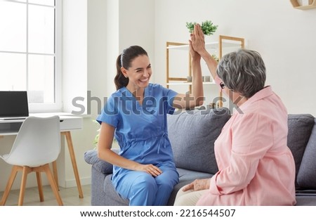 Cheerful positive female nurse giving high five to her senior female patient during visit. Caregiver is sitting on sofa with retired patient and congratulates her on successful outcome of treatment.