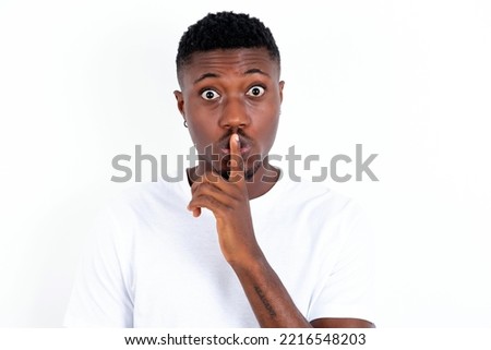 young handsome man wearing white T-shirt over white background makes hush gesture, asks be quiet. Don't tell my secret or not speak too loud, please!