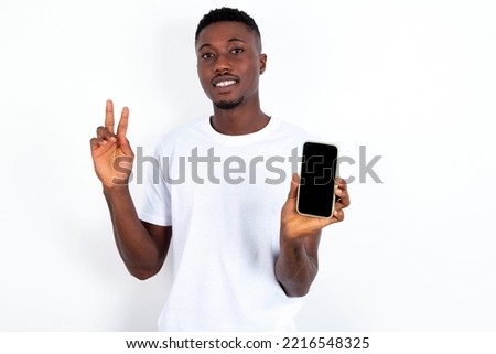 young handsome man wearing white T-shirt over white background holding modern device showing v-sign