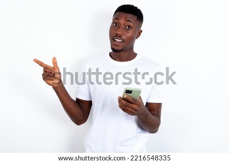 Astonished young handsome man wearing white T-shirt over white background holding her telephone and pointing with finger aside at empty copy space