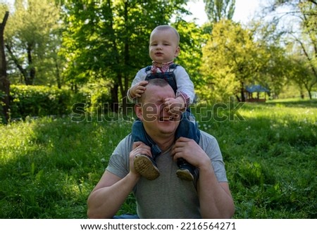 A funny photo where dad holds his little son on his shoulders, dressed in Ukrainian vyshyvanka and denim overalls in the park, the kid bending over pulls dad by the nose, dad is laughing