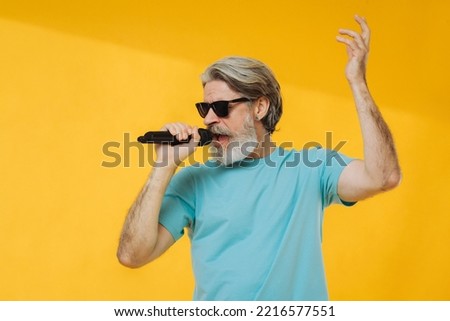 Photo of gray-haired senior man singing, wearing glasses, blue t-shirt, isolated on yellow background