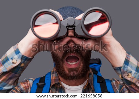 Wow! Surprised young bearded man carrying backpack and looking through binoculars while standing against grey background