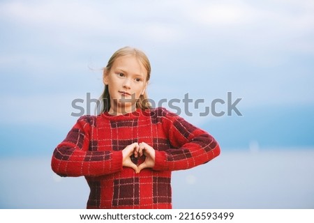 Outdoor portrait of cute little girl showing heart shaped love sing, wearing red pullover, posing next to water