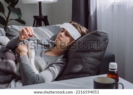 Woman under plaid feel sick unwell. Unhealthy young female measuring body temperature with electronic thermometer, suffer from flu lying on sofa at home, seasonal contagious grippe illness concept Royalty-Free Stock Photo #2216594051