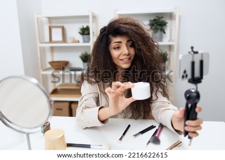 Smiling tanned handsome curly Latin beauty blogger in linen shirt present facial cream in home white interior. Copy space Mockup Banner. Influencer record video review blog using smartphone Royalty-Free Stock Photo #2216622615