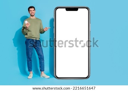 Full size photo of cute brunet millennial guy hold telephone index promo wear shirt jeans shoes isolated on blue background