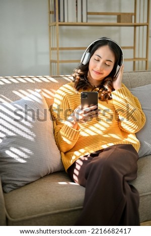 Portrait, Relaxed and clam young Asian female in casual clothes chilling on a comfortable couch in the living room, using her smartphone and listening to music through her headphones.