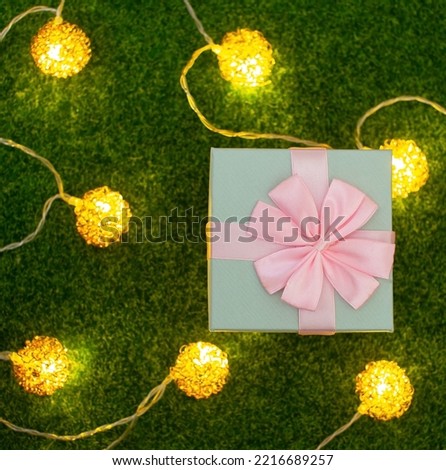 A blue gift box tied with a pink bow on a green fake grass with decorative lights surrounding it.