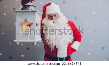 Portrait of man in Santa suit holding lantern and smiling looking at camera on starry grey background. Holidays and festivals concept.