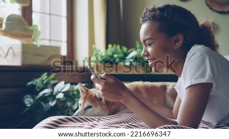Pretty girl student is using smartphone touching screen and laughing lying on bed at home with cute pet dog, animal is enjoying care and love. Technology and animals concept. Royalty-Free Stock Photo #2216696347