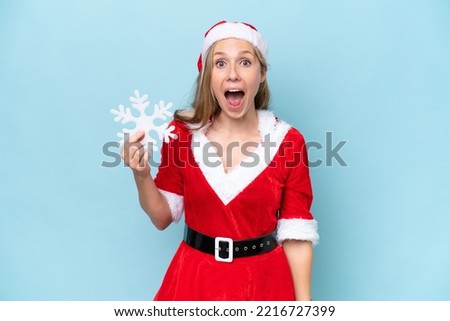 Young caucasian woman dressed as Mama Claus holding a snowflake isolated on blue background with surprise and shocked facial expression