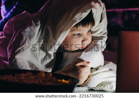 Happy kid son laughing eat popcorn remote control watching funny comedy tv show sitting on sofa having fun viewing video on laptop in evening at home. Teenager gen z boy under blanket at Movie night