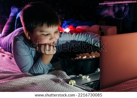 Cheerful teen boy using laptop at home watching funny movie cartoon eat pop corn, browsing internet on sofa in living room. Smiling kid looking at screen having fun enjoying weekend night vacation.
