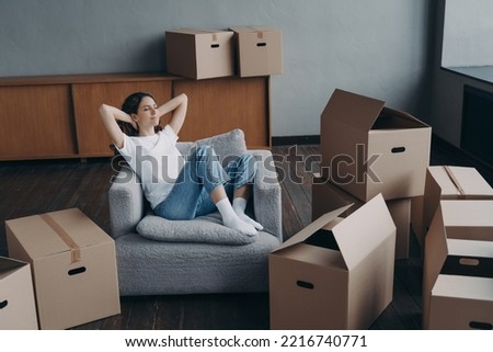 Relaxed woman sitting on armchair in empty house apartment with hands behind head, happy young female homeowner tenant relaxing during packing boxes on moving day to new home. Easy relocation concept. Royalty-Free Stock Photo #2216740771