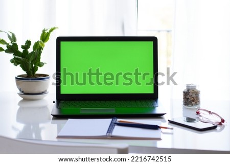 Non-people image of desk setup, work environment. Selective focus of laptop with green screen, notebook, pen, smartphone, glasses, flower on the table in front of window. Concept of distance education