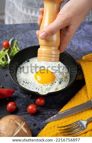 female hands using a wooden mill for spices prays peppers on fried eggs in a cast-iron skillet