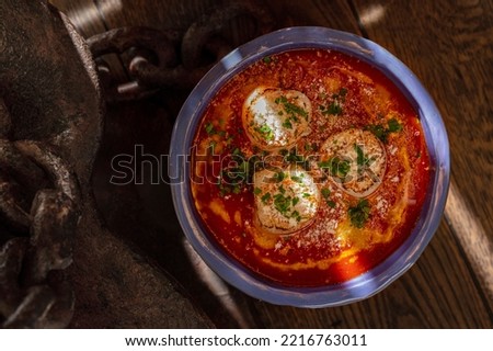 Sea scallop fried in white wine with tomato sauce and wig, finely chopped cilantro. The food lies in a ceramic, blue plate with high sides. The plate stands on a dark, wooden background.