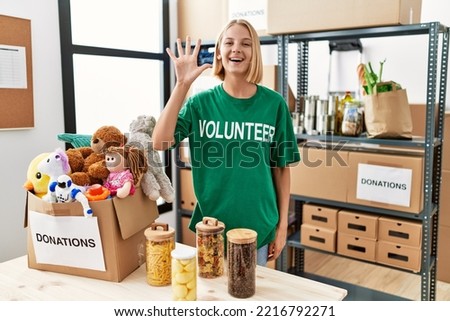 Young caucasian woman wearing volunteer t shirt at donations stand showing and pointing up with fingers number five while smiling confident and happy. 