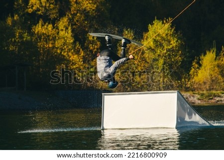 A wakeboarder in a wetsuit performs a trick on a lake against the background of an autumn forest. A lake in the mountains with a wakeboarder. Wakeboard in the mountains. Recreation