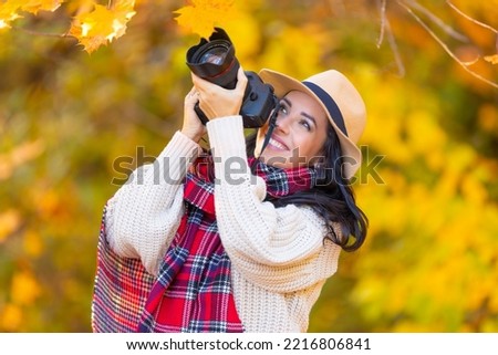 Smiling good-looking female photographer in a hat and stylish autumn clothes takes pictures outside on a sunny fall day.