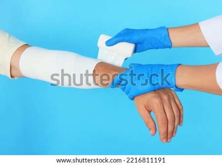Doctor applying medical bandage onto patient's arm on light blue background, closeup
