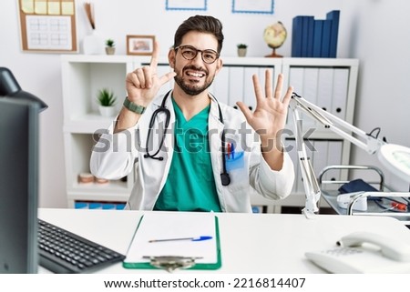 Young man with beard wearing doctor uniform and stethoscope at the clinic showing and pointing up with fingers number seven while smiling confident and happy. 