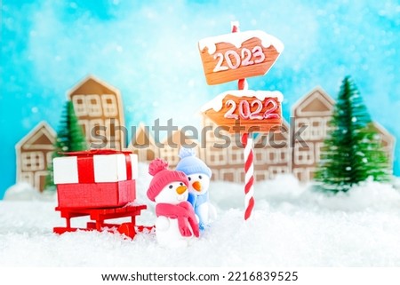 Two cute plasticine snowmen with sleds and gifts in a toy town. Wooden direction signs 2023 and 2022. Waiting for the New Year 2023, Christmas card.
