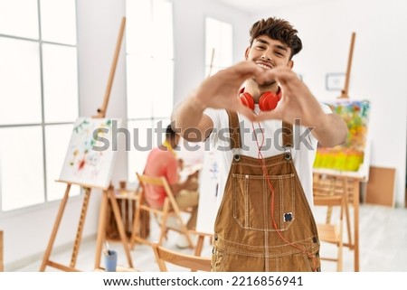 Young hispanic man at art studio smiling in love doing heart symbol shape with hands. romantic concept. 
