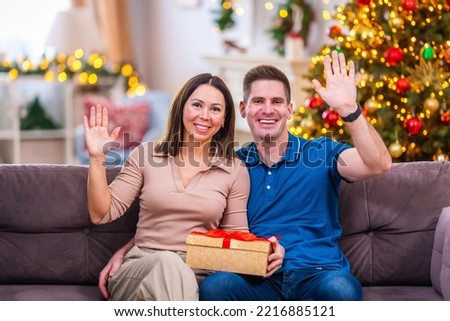 young man gives gift for holiday to his beloved woman and is greeted. An attractive girl and guy smiling happily and waving their hands happily, sitting on sofa in living room decorated for Christmas.