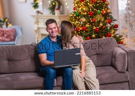 young married couple uses a laptop while sitting on a sofa in a decorated living room, chatting with friends and shopping online at Christmas sale. girl and young man hug, kiss and have fun together.