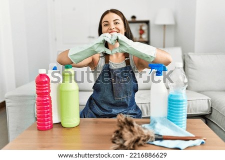 Young brunette woman wearing cleaner apron and gloves cleaning at home smiling in love doing heart symbol shape with hands. romantic concept. 