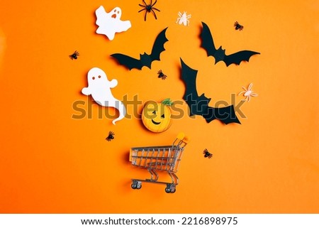 Shopping trolley with decorative pumpkin, ghosts, bats and spiders on orange background. Flat lay Halloween sale concept. 