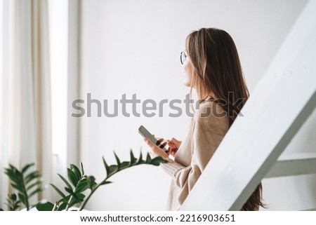Smiling woman with long hair in beige cardigan drinking coffe and using mobile phone at home