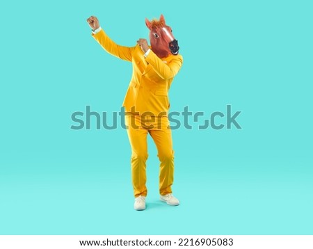Full length portrait of funny playful showman in yellow suit and rubber pink horse mask dancing on turquoise background. Banner for advertisement, marketing with funky man. He is rising his hands up. Royalty-Free Stock Photo #2216905083