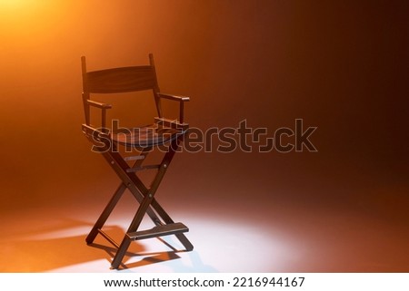 The director's chair stands in a beam of light with an orange backlight and smoke. Place for text. Free chair. Concept of selection and casting. Shadow and light. Royalty-Free Stock Photo #2216944167