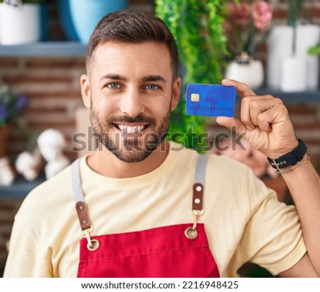 Handsome hispanic man working at florist shop holding credit card looking positive and happy standing and smiling with a confident smile showing teeth 
