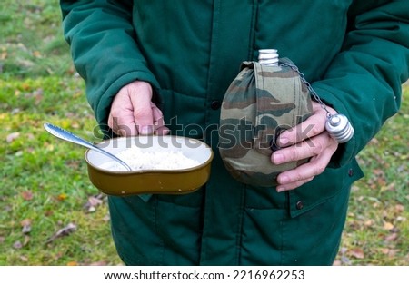A man holds in his hands a military flask for water and an army bowler hat with porridge and a spoon close-up