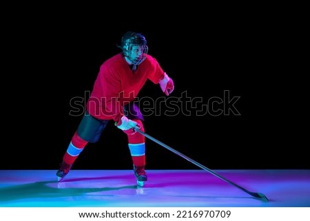 Junior ice hockey player in sports uniform and protective equipment in action over dark background in neon light. Sport, power, challenges, achievement, goals. Young boy playing hockey