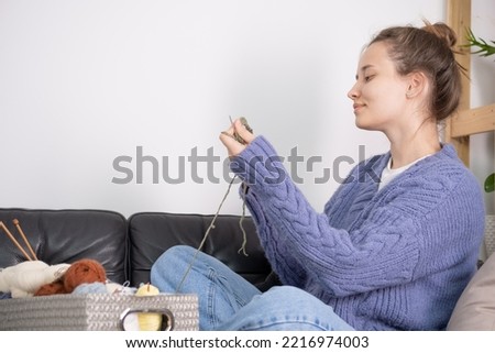 A young reaper knits a hat. The woman is dressed in a sweater in the color of peri peri. The process of knitting a hat on knitting needles. Close angle