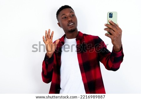 young handsome man wearing red plaid shirt over blue background holds modern mobile phone and makes video call waves palm in hello gesture. People modern technology concept
