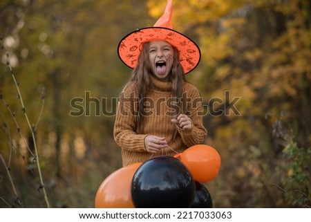 Halloween kids, portrait of a girl in a sorcerer hat with balloons showing tongue, girl having fun on Halloween outdoors in the forest, happy halloween. High quality photo