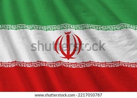 Iran flag with big folds waving close up under the studio light indoors. The official symbols and colors in fabric banner
