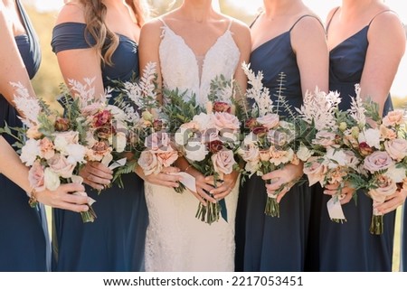 Bride and bridesmaids holding bouquets. Bridesmaids are wearing navy blue dresses. Bouquet has roses and wildflowers.  Royalty-Free Stock Photo #2217053451