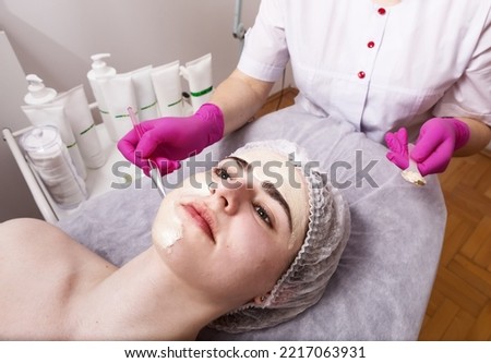 Hands of a beautician in pink medical gloves apply a gold cosmetic mask on the face of a beautiful girl with piercings in a beauty salon, horizontal, close-up
