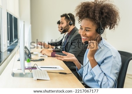 portrait of smiling mixed race female call center or help desk or telephone support agent working at call center workplace wearing headset with blurred background of male agent Royalty-Free Stock Photo #2217067177